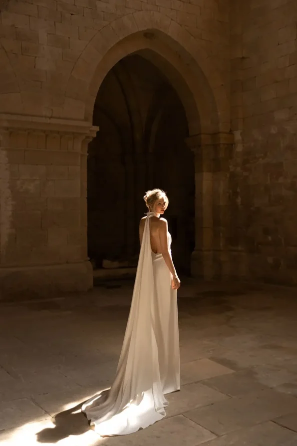 A woman in a long white Rouge by Eva Lendel dress with a cape stands in a sunlit stone hall with arched doorways.
