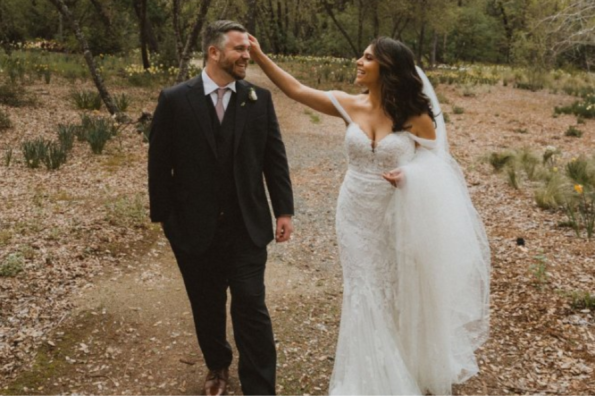 A bride in a white wedding dress, inspired by 2025-2026 wedding dress trends, gently touches the groom's hair as they walk together on a path in an outdoor, wooded San Francisco Bay Area setting.