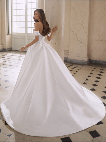 Etoile woman wearing the FREYA by Etoile, an off-the-shoulder white ball gown with a long train, stands in a grand marble room with a checkered floor, glancing over her shoulder.