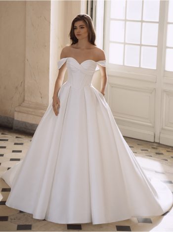 A woman poses indoors in the FREYA by Etoile off-the-shoulder white ball gown wedding dress, featuring a fitted bodice and full skirt, standing before large windows and marble walls.