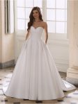 A woman poses indoors in the FREYA by Etoile off-the-shoulder white ball gown wedding dress, featuring a fitted bodice and full skirt, standing before large windows and marble walls.