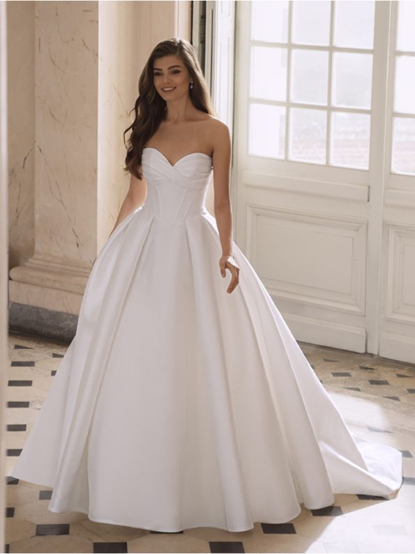 A woman poses indoors in the FREYA by Etoile strapless white ball gown, featuring a fitted bodice and full skirt, standing before tall windows and marble columns.