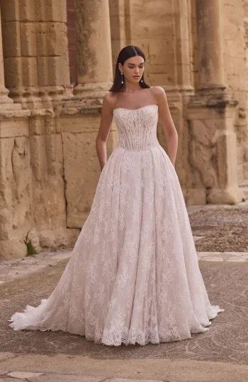 A woman stands outdoors in front of a stone building with columns, wearing product 63005A—a strapless lace wedding gown.