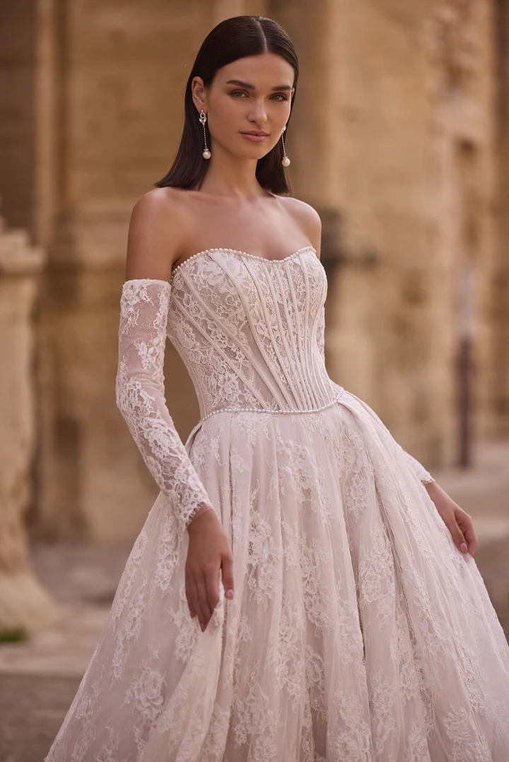A woman in the 63005A off-the-shoulder lace wedding dress with long sleeves and a fitted bodice stands outdoors against a stone building. A woman in the 63005A off-the-shoulder lace wedding dress with long sleeves and a fitted bodice stands outdoors against a stone building.