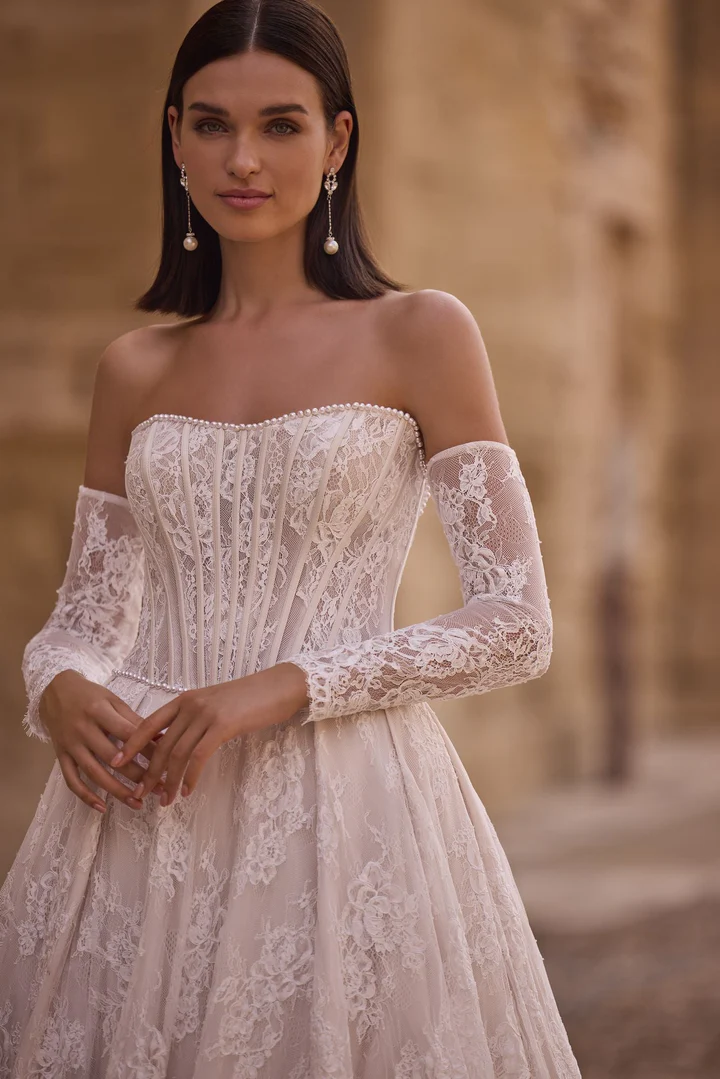 A woman in the 63005A off-the-shoulder lace wedding dress with sheer long sleeves and pearl details stands outdoors. A woman in the 63005A off-the-shoulder lace wedding dress with sheer long sleeves and pearl details stands outdoors.