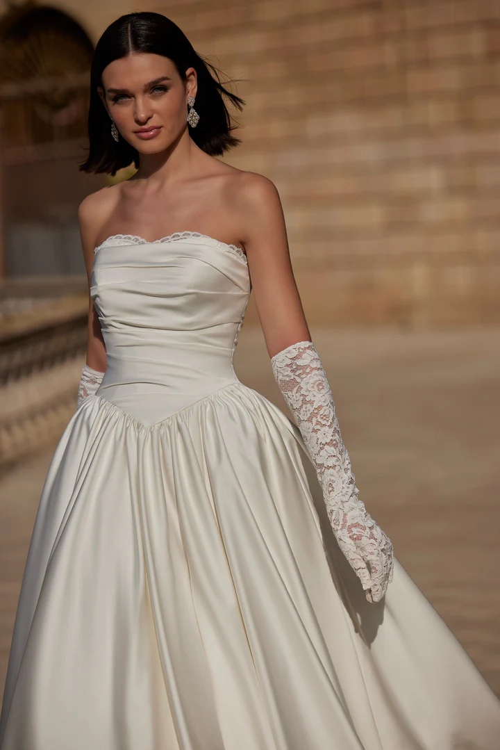 A woman wearing product 63114, a strapless white satin gown with long lace gloves, stands outdoors in front of a stone wall. A woman wearing product 63114, a strapless white satin gown with long lace gloves, stands outdoors in front of a stone wall.