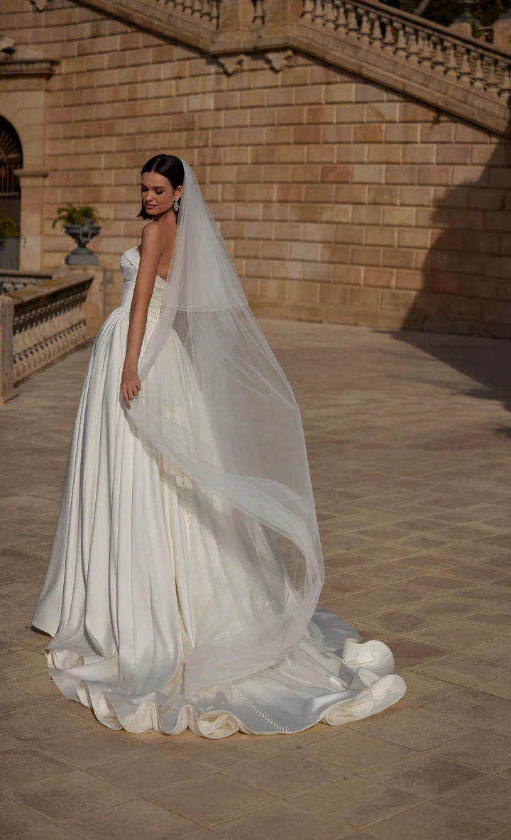 A bride in style 63114, a strapless white gown with a long train and veil, stands outdoors on a stone terrace looking over her shoulder. A bride in style 63114, a strapless white gown with a long train and veil, stands outdoors on a stone terrace looking over her shoulder.