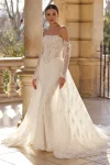 A woman is outdoors wearing wedding gown 63115—an off-the-shoulder lace dress with sheer long sleeves, a fitted bodice, and a long train.