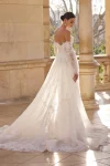 A woman is outdoors wearing wedding gown 63115—an off-the-shoulder lace dress with sheer long sleeves, a fitted bodice, and a long train.