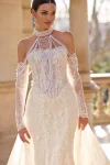 A woman is outdoors wearing wedding gown 63115—an off-the-shoulder lace dress with sheer long sleeves, a fitted bodice, and a long train.