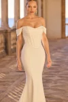 A woman stands on a stone terrace wearing product 63156, an off-the-shoulder, form-fitting white gown with a fitted bodice and slight train, looking directly at the camera.