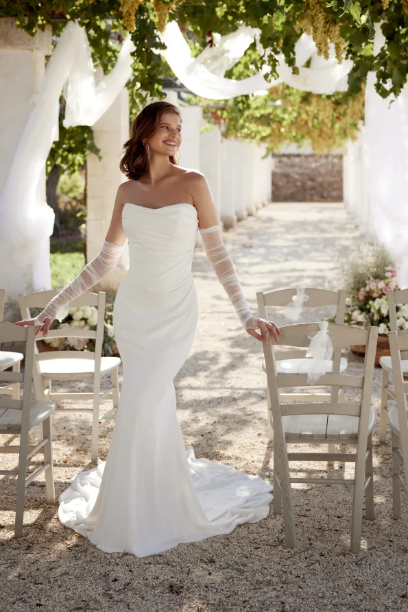 A woman wearing the 66388 by Lillian West wedding dress stands outdoors between rows of wooden chairs, surrounded by lush greenery and white decorations.