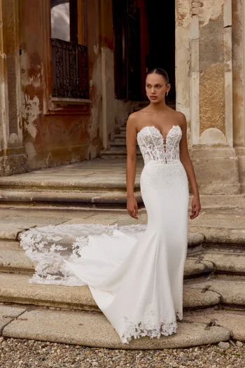 A woman stands on stone steps before an old building, wearing the HOPE/88353 by Justin Alexander: a strapless, fitted white wedding gown with a sheer lace bodice and long lace train.