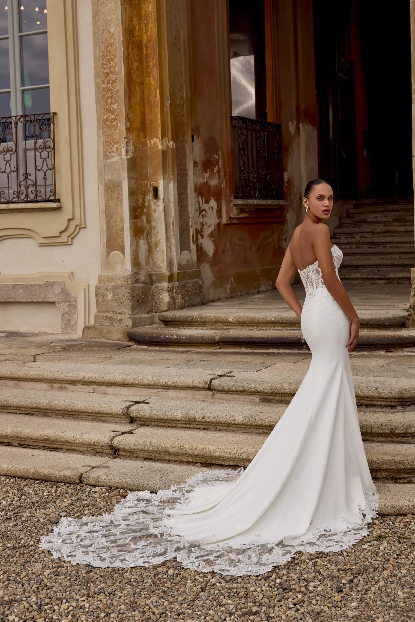 A woman in the HOPE/88353 by Justin Alexander, a strapless white wedding gown with a lace train, stands on stone steps before an old building. A woman in the HOPE/88353 by Justin Alexander, a strapless white wedding gown with a lace train, stands on stone steps before an old building.