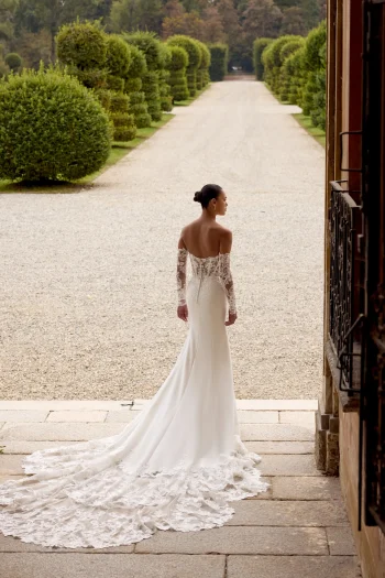 A woman stands facing away outdoors near a gravel path, wearing the HOPE/88353 by Justin Alexander—a white, off-the-shoulder wedding dress with a long lace train.
