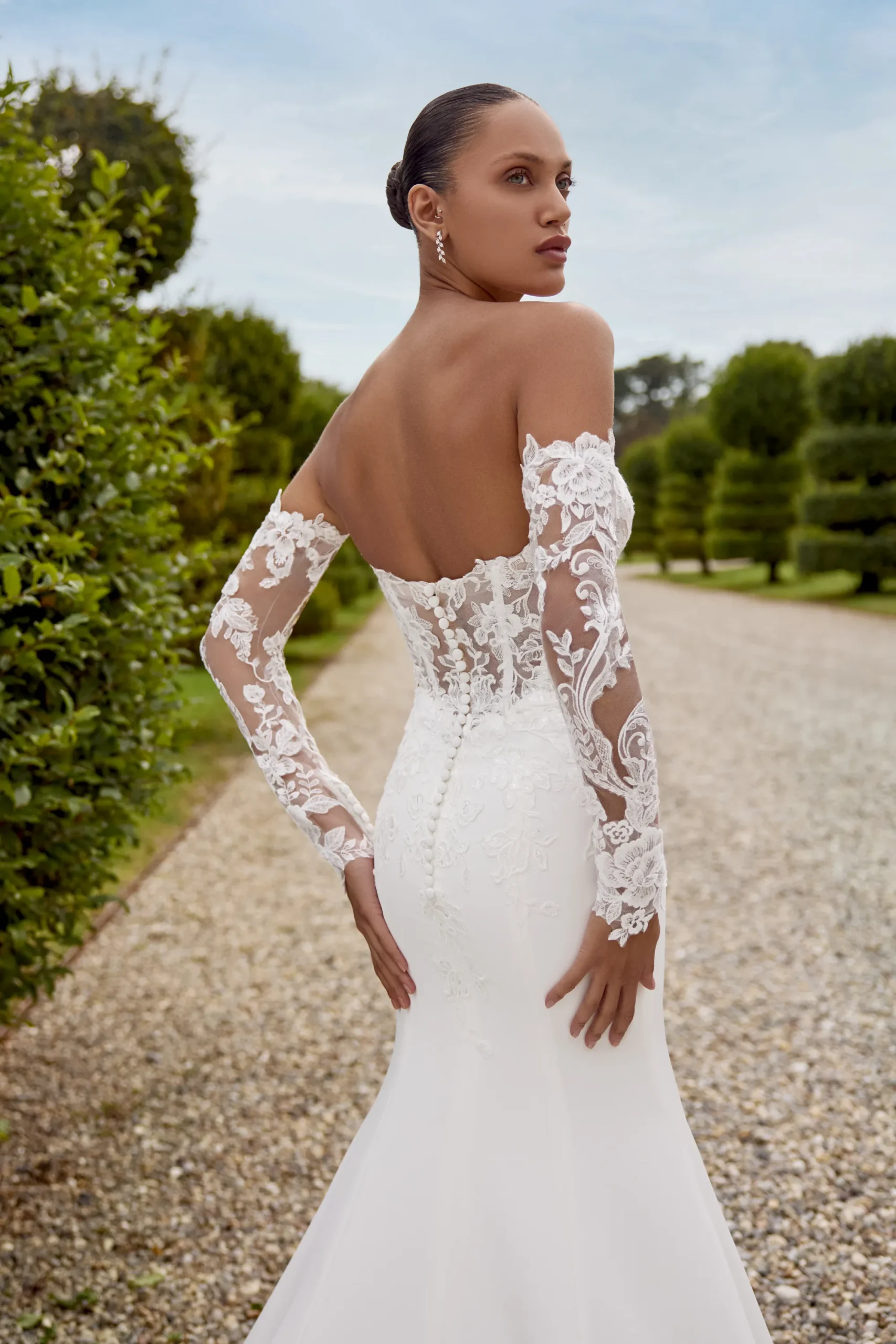 Wearing the HOPE/88353 by Justin Alexander, an off-the-shoulder lace wedding dress, a woman stands on a gravel path bordered by greenery, glancing gracefully over her shoulder. Wearing the HOPE/88353 by Justin Alexander, an off-the-shoulder lace wedding dress, a woman stands on a gravel path bordered by greenery, glancing gracefully over her shoulder.