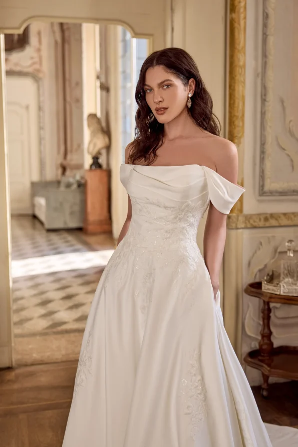 A woman wears the LARISSA/88491 by Justin Alexander—an off-the-shoulder bridal gown—while standing indoors, surrounded by ornate walls and checkered flooring.