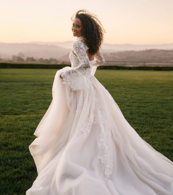 Wearing the Allure A1408 Strapless Sequin Lace Ball Gown Wedding Dress, a woman stands on the grass at sunset, gazing back over her shoulder with a gentle smile.