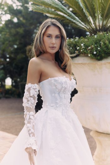 A woman wearing the ARTEMIS wedding gown (style AW2583), an off-shoulder white lace dress, stands outdoors near a large planter filled with greenery, gazing at the camera.