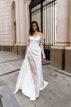 A woman wearing the BALENSOI white off-the-shoulder wedding dress with lace sleeves and a thigh-high slit walks on a paved city street near a black iron gate and stone columns.