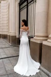 A woman wearing the BALENSOI white off-the-shoulder wedding dress with lace sleeves and a thigh-high slit walks on a paved city street near a black iron gate and stone columns.