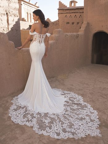 A bride stands on a sunlit adobe terrace, facing away in the TAMMY by Enzoani off-shoulder fitted white gown with a long lace train.