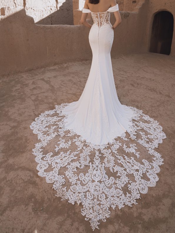 A woman in the off-shoulder, fitted TAMMY by Enzoani bridal gown with a long lace train stands on sandy ground near an adobe wall.