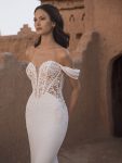 A woman in the TAMMY by Enzoani off-the-shoulder, fitted bridal gown with lace detailing stands before an adobe-style building.