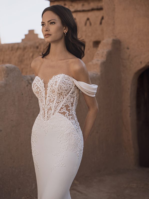 A woman stands before an adobe-style building in the TAMMY by Enzoani—a white, off-the-shoulder lace bridal gown with a fitted silhouette and sheer bodice.