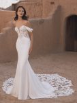 A woman in the TAMMY by Enzoani off-the-shoulder, fitted bridal gown with lace detailing stands before an adobe-style building.
