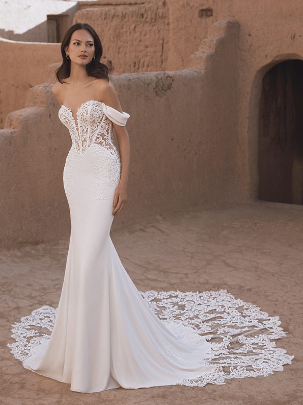 A woman in the TAMMY by Enzoani off-the-shoulder, fitted bridal gown with lace detailing stands before an adobe-style building.
