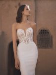 A woman in the TAMMY by Enzoani off-the-shoulder, fitted bridal gown with lace detailing stands before an adobe-style building.
