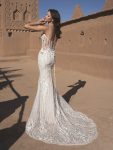 A woman in a fitted white lace THAO by Enzoani bridal gown with thin straps stands outside against an adobe wall under a clear sky.