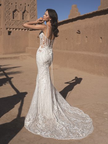 A woman wearing the THAO by Enzoani fitted bridal gown with lace details and a long train stands outdoors near adobe-style walls under a clear sky.