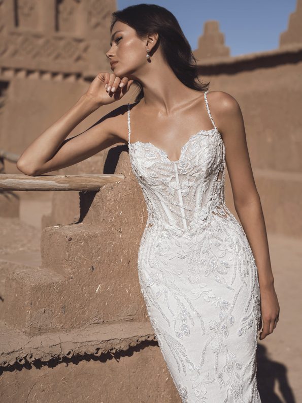 A woman in a fitted THAO by Enzoani white lace bridal gown with thin straps leans against an adobe wall beneath a clear blue sky.