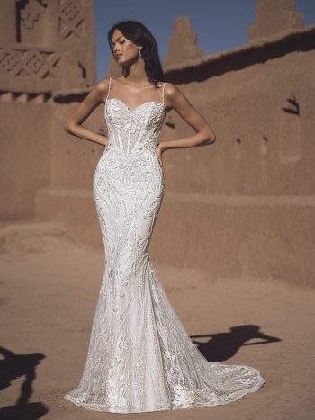 A woman in a fitted white lace THAO by Enzoani bridal gown with thin straps stands outside against an adobe wall under a clear sky.