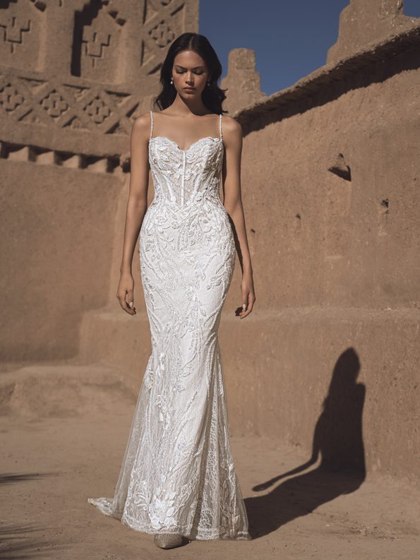 A bride in a fitted, white lace THAO by Enzoani gown with thin straps stands outdoors beside a textured sand-colored wall under a clear blue sky.