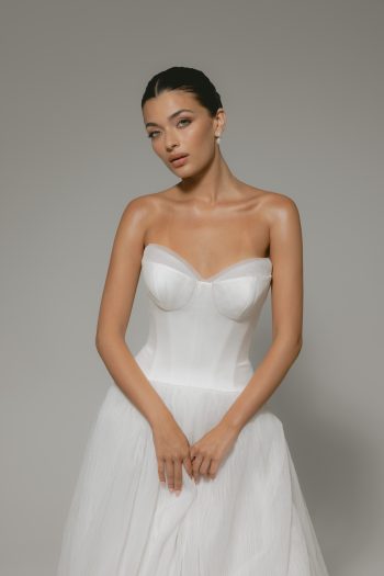 A woman in a strapless white CASSIA/AW2614 dress stands against a plain gray background, hands gently clasped in front of her.