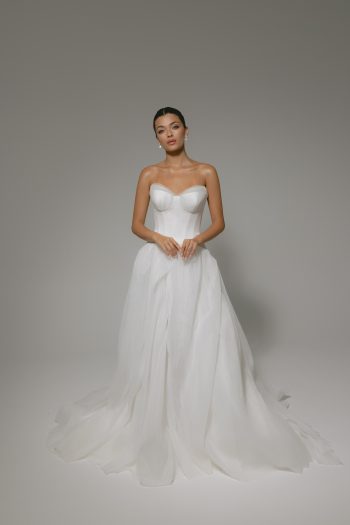 A woman poses against a neutral background, wearing the elegant CASSIA/AW2614—an exquisite strapless, white, floor-length wedding gown with a flowing skirt.