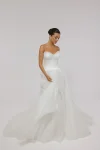 A woman in a strapless white CASSIA/AW2614 dress stands against a plain gray background, hands gently clasped in front of her.