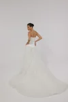 A woman in a strapless white CASSIA/AW2614 dress stands against a plain gray background, hands gently clasped in front of her.
