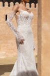 A woman in the white lace CINDY/JC2597 off-the-shoulder wedding dress with long sheer lace sleeves stands before a beige textured background.