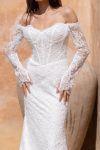 A woman in the white lace CINDY/JC2597 off-the-shoulder wedding dress with long sheer lace sleeves stands before a beige textured background.