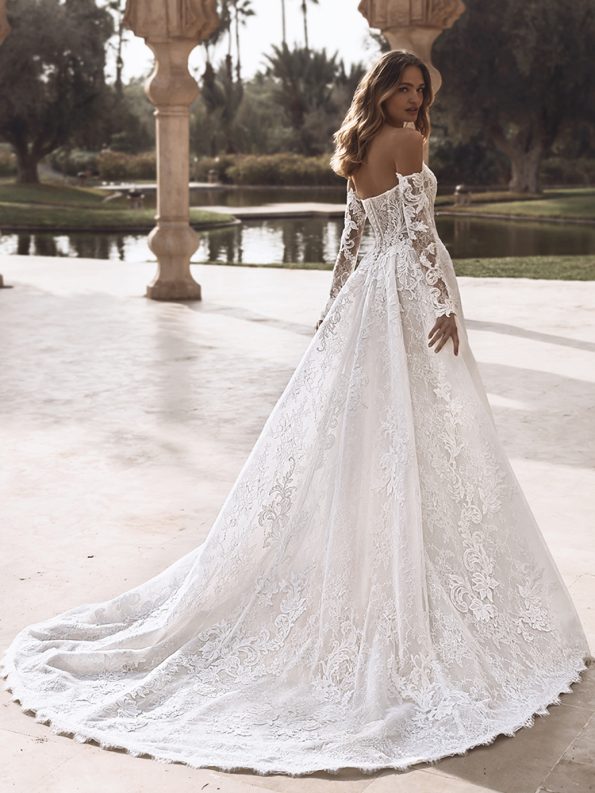 A woman stands outdoors by a pond, wearing the VANESSA by Enzoani—a long-sleeved, off-the-shoulder lace wedding dress with a sweeping train.