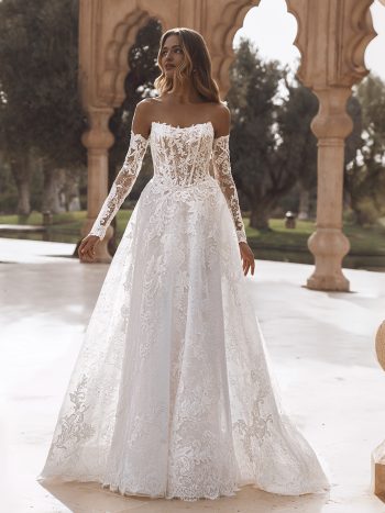 A woman stands outdoors in VANESSA by Enzoani, a white off-the-shoulder lace wedding dress with long sleeves and a full skirt, framed by stone arches and trees.
