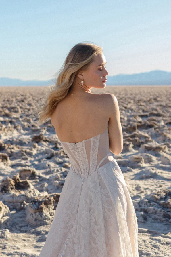A woman in the strapless white JULIETTE/F380 by Allure dress stands in a rocky desert landscape, gazing sideways with distant mountains, radiating effortless allure.