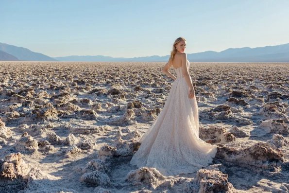 A woman in a white lace dress stands on a rocky, salt-strewn landscape with mountains behind her and a clear sky above, radiating the timeless elegance of JULIETTE/F380 by Allure.