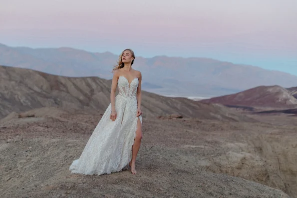A woman in the JULIETTE/F380 by Allure strapless white wedding dress stands barefoot on rocky terrain, with mountains behind her beneath a pastel sky.