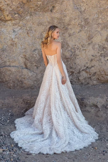 A woman wearing the JULIETTE/F380 by Allure strapless white lace gown stands outdoors before a rocky, earth-toned wall, facing slightly away from the camera.
