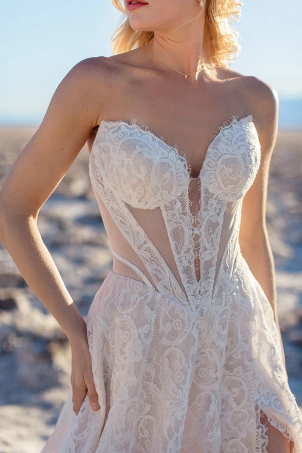 A woman in the JULIETTE/F380 by Allure, a strapless lace wedding dress with a deep neckline, stands outdoors on sunlit sand, radiating effortless allure.